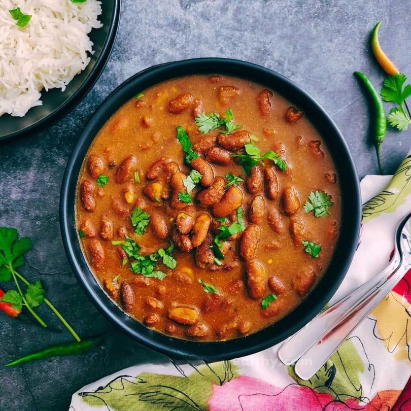 A black bowl of kidney bean curry with cilantro, rice, chilies, and a floral napkin.