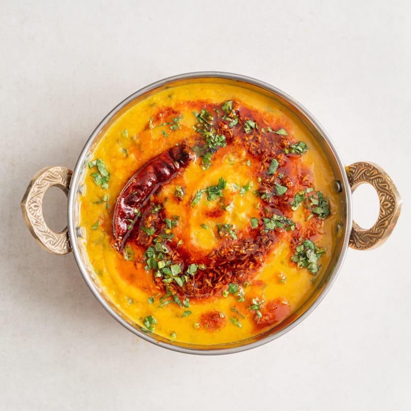 Overhead view of a golden dal in a metal bowl, garnished with red oil, chili, and cilantro.