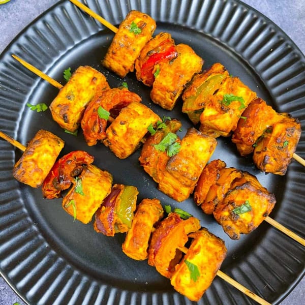 Three skewers of paneer tikka, with cheese cubes and bell peppers, on a black plate.